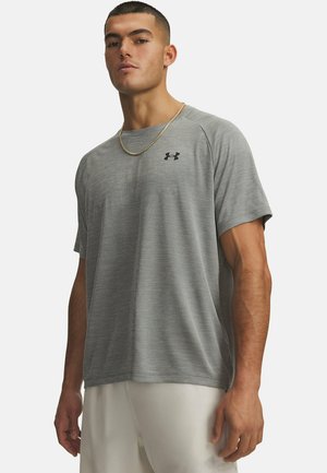 Man staat met een grijs Under Armour T-shirt met korte mouwen, witte shorts en een gouden ketting tegen een effen achtergrond.