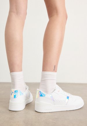 White sneaker with reflective iridescent accents, perforated design, and a textured sole, worn with white crew socks.