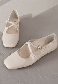 Fehér bőrből készült balettcipő négyzet alakú orral, keresztpánttal és ezüst szegecses díszítéssel a felső részen. Puha belső bélés. Lapos sarok.