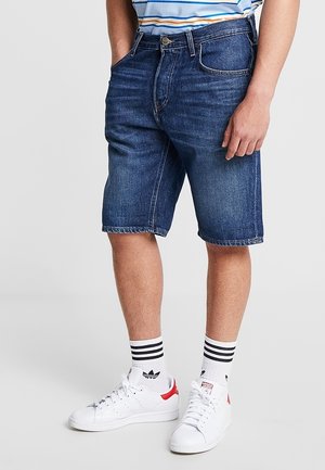 Persoon die blauwe denimshorts draagt, witte sneakers met rode accenten en witte sokken met zwarte strepen, staand met één hand in de zak.