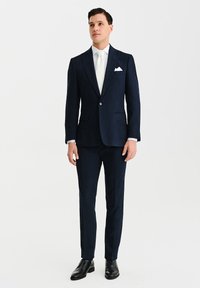 Granatowy garnitur o dopasowanym kroju, z szpiczastymi klapami i matową teksturą. W komplecie z białą koszulą, krawatem i poszetką, noszony z czarnymi butami.