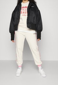 Svart vadderad bomberjacka, krämfärgad sweatshirt med röd tryck, krämfärgade joggers med röda sidosträck, vita sneakers och strumpor med rosa ränder.