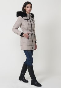 Abrigo de plumas beige con un ribete de piel sintética negra, cintura entallada y bolsillos con cremallera. Combinado con unos vaqueros azul oscuro y botas negras hasta la rodilla.