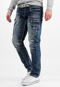 Jeans in denim blu con una vestibilità slim, dotati di numerose tasche con zip e cuciture. Indossati con sneakers bianche e una felpa grigia.