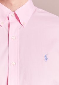 Ljust rosa skjorta med knappar, tillverkad av bomull, med klassisk krage och broderad blå logotyp på vänster sida. Stora, vita knappar.