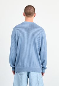 Suéter azul de punto con textura acanalada y mangas largas. La vista posterior muestra un cuello redondo y un corte relajado. Combinado con pantalones cortos azul claro.
