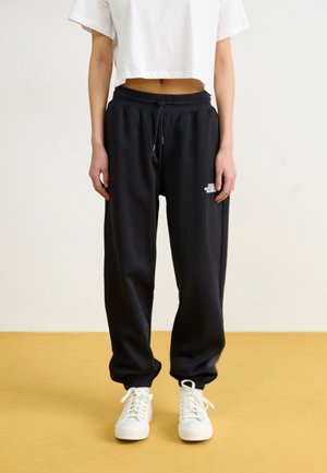 Person wearing black North Face sweatpants, white cropped t-shirt, and white sneakers, standing on a yellow floor against a white wall.