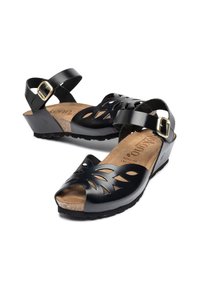 Sandales en cuir noir avec motif floral découpé, bride de cheville réglable avec boucle dorée, semelle en liège et talon compensé.