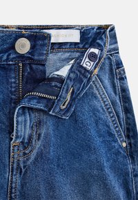 Vaqueros de mezclilla en azul oscuro con cierre de cremallera y botón. Cuenta con una etiqueta de marca y detalles de costura en la cintura y los bolsillos.