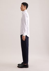 Wit overhemd met kraag en dunne verticale strepen, gecombineerd met marineblauwe broek en zwarte nette schoenen, van opzij getoond tegen een neutrale achtergrond.