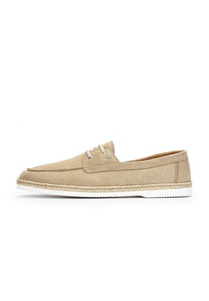 Zapato de ante beige con un diseño plano, suela de goma blanca y punta suave y redondeada. Presenta ojales de metal y un detalle de yute tejido alrededor de la suela.