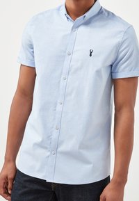 Camisa de manga corta de color azul claro, hecha de una mezcla de algodón. Tiene un cuello tipo button-down, una tapeta central con botones y un pequeño logotipo de ciervo bordado.