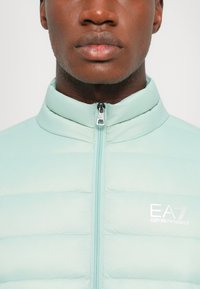 Jachetă puffer de culoare verde mentă deschisă, cu guler înalt, închidere frontală cu fermoar, cusături orizontale și logo brodat EA7 pe partea stângă.