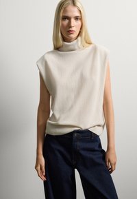 Beige ärmelloser Rollkragenpullover aus weichem Strickmaterial mit lockerer Passform. Kombiniert mit dunkelblauen hoch taillierten Jeans, die Standardtaschen haben.