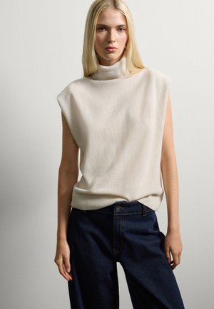 Maglione senza maniche color beige in morbido materiale a maglia, con una vestibilità rilassata. Abbinato a jeans a vita alta di colore blu scuro, dotati di tasche standard.