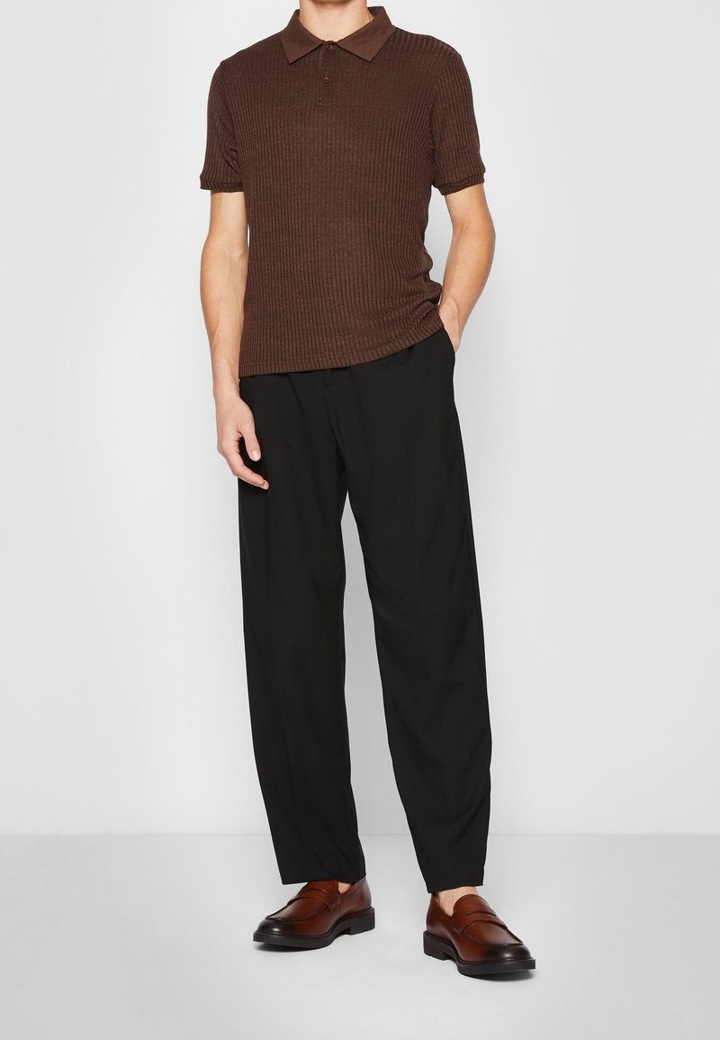 Polo marron côtelé à manches courtes, associé à un pantalon noir ample et à des mocassins marron foncé. Design simple et ajusté.