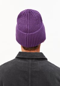 Lila gestrickte Mütze mit vertikaler Rippstruktur. Wird auf dem Kopf einer Person getragen, mit einem strukturierten dunklen Hemd, das im Vordergrund sichtbar ist.