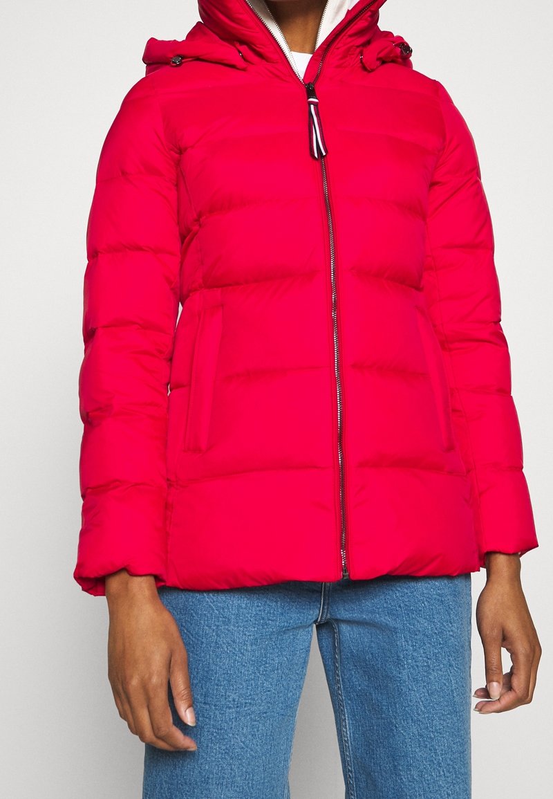 Veste matelassée rouge avec fermeture éclair à l'avant, poches latérales et capuche, portée avec un jean bleu sur une personne devant un fond uni.