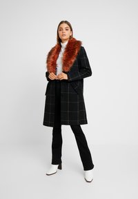 Kvinne iført en svart rutete frakk med stor rustrød pelskrage, hvit rullekragegenser, svarte bukser og hvite ankelstøvler, som står foran en hvit bakgrunn.