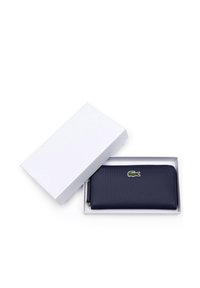 Portefeuille zippé navy avec surface texturée, présentant un petit logo Lacoste vert et rouge, présenté dans une boîte cadeau blanche.