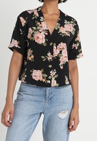 Blusa nera a fiori con maniche corte, decorata con rose rosa e foglie verdi, scollatura a V e chiusura con bottoni, abbinata a jeans strappati blu chiaro.