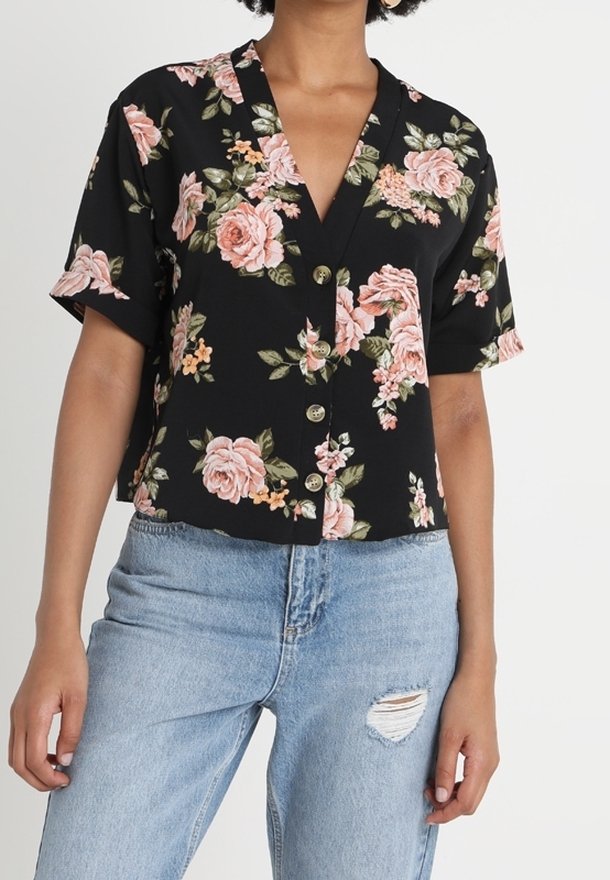 Blusa nera a fiori con maniche corte, decorata con rose rosa e foglie verdi, scollatura a V e chiusura con bottoni, abbinata a jeans strappati blu chiaro.