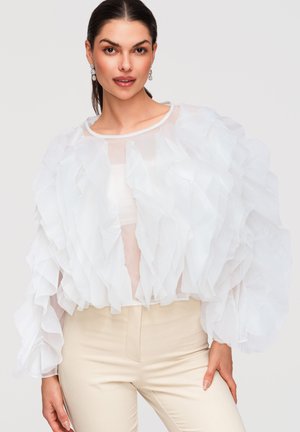 Femme vêtue d'un chemisier blanc transparent à volants et d'un pantalon beige, posant avec la main sur la hanche sur un fond uni.