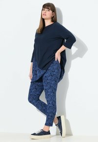 Donna con una maglietta a maniche lunghe blu navy, leggings blu con motivo e sneakers blu navy, in piedi contro un muro bianco, con una mano sui fianchi.
