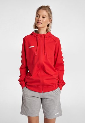 Rode zip-up hoodie van zacht materiaal met wit logo en chevron strepen op de mouwen. Gemaakt in combinatie met grijze shorts met logo-detail.