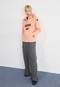 Jachetă impermeabilă roz cu un buzunar frontal și logo, purtată cu pantaloni largi vrac cu dungi gri și pantofi negri. Design simplu și croială spațioasă.