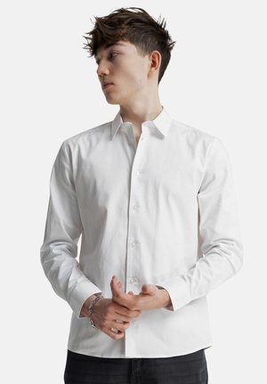 Młody mężczyzna o krótkich brązowych włosach, ubrany w białą koszulę na guziki i srebrną bransoletkę, patrzący w lewo na tle jednolitego tła.