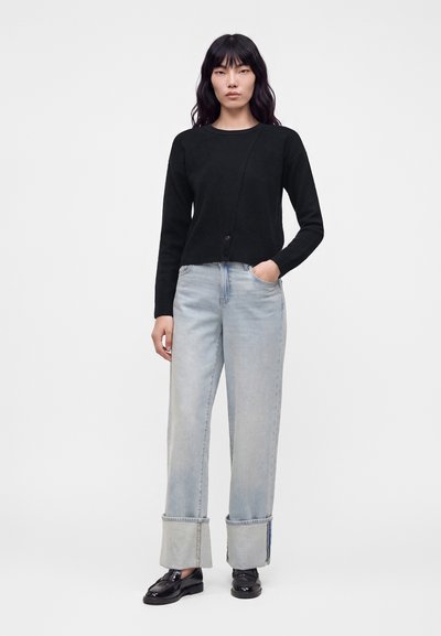 Femme debout portant un haut noir à manches longues, un jean bleu clair à jambes larges avec revers, et des mocassins noirs brillants sur un fond uni.