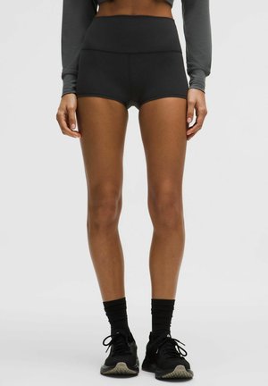 Kvinde iført sorte, højtaljede træningsshorts, sorte crew-sokker og sorte løbesko, stående mod en enkel, lys baggrund.