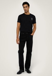 Camiseta de algodón negra con un pequeño gráfico en el pecho, combinada con jeans negros. El modelo está de pie con botas negras contra un fondo neutro.