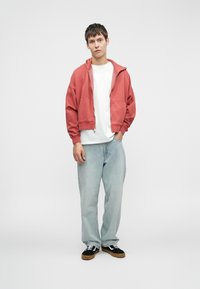 Κόκκινο φούτερ με φερμουάρ, λευκή μπλούζα, ανοιχτό μπλε τζιν και μαύρα αθλητικά παπούτσια με καφέ σόλες. Το μοντέλο στέκεται με τα χέρια στις τσέπες σε ένα απλό φόντο.