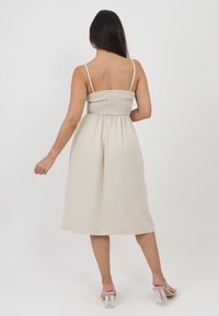Ärmelloses, hellbeiges Kleid mit einem gerafften Oberteil und einem fließenden Rock, aus weichem Material gefertigt. Verfügt über dünne Träger und eine Midi-Länge.