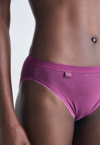 Gros plan sur le torse d'une personne à la peau foncée portant des sous-vêtements en coton violet avec une étiquette "Sloggi" sur la ceinture.