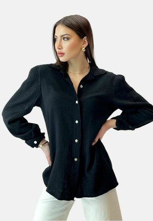 Vrouw met steil bruin haar draagt een zwarte, gestructureerde blouse met knopen en witte broek, poseert met handen in de zij en kijkt naar links.