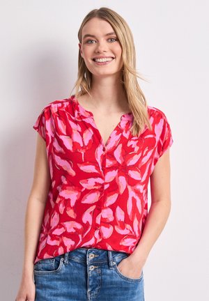 Smilende kvinde med langt blond hår iført en rød kortærmet bluse med lyserøde bladmotiver og blå jeans, stående med den ene hånd i lommen.