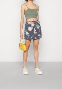 Žena ve zeleném croptopu, námořnických květovaných šortkách, bílých platformových teniskách, držící malou žlutou kabelku na šedé podlaze a bílém pozadí.