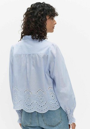 Femme aux cheveux bouclés portant un chemisier à rayures bleues et blanches avec broderie ajourée et ourlet festonné, associé à un jean bleu clair.