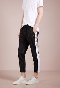 Schwarze schmal zulaufende Sweatpants mit weißen Seitenstreifen und gelben Sternakzenten, aus einem glatten Stoff gefertigt. Lässige Passform, kombiniert mit weißen Sneakers.