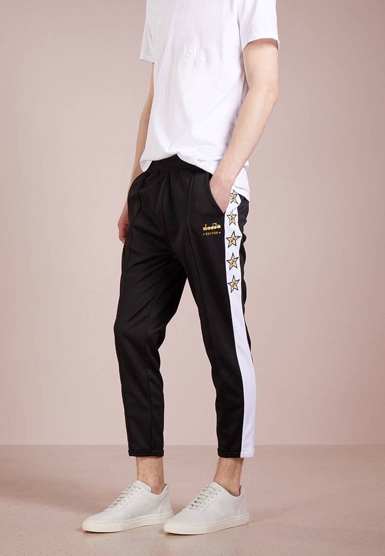 Schwarze schmal zulaufende Sweatpants mit weißen Seitenstreifen und gelben Sternakzenten, aus einem glatten Stoff gefertigt. Lässige Passform, kombiniert mit weißen Sneakers.