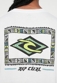 Bijela majica sa velikim crno-zelenim logotipom u središtu, okružena šarenim geometrijskim uzorakom i tekstom "RIP CURL."