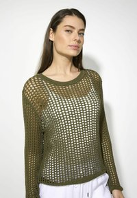 Olivgrüner, locker gestrickter Pullover mit weitem Ausschnitt, langen Ärmeln und einem offenen Maschendesign, getragen über einem weißen Oberteil.