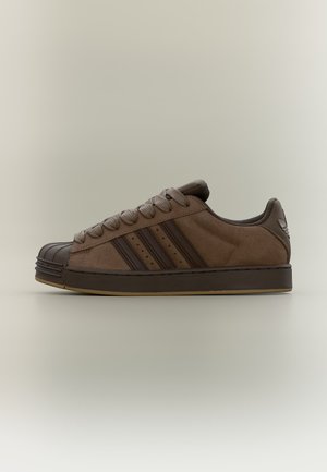 Smeđi niskoprofilni tenisica s tamnosmeđim prugama, gumeni zaštitni vrh, vezice i ravni potplat na svijetloj neutralnoj pozadini.