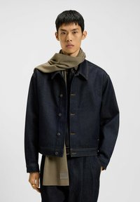 Mørk denimjakke med brune sømcirkler, som har en klassisk krave og knaplukning. Bæres over en mørk skjorte med et olivengrønt tørklæde.