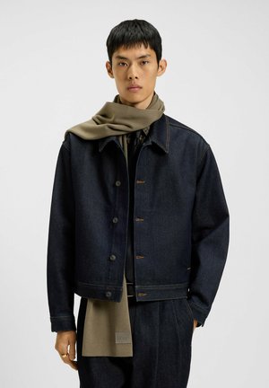 Dark denim jacket with brown stitching, featuring a classic collar and button closure. Worn over a dark shirt with an olive green scarf.
