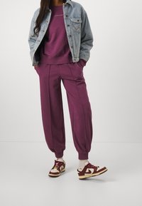 Violetti ylisuurit collegehousut, joissa on kapeneva resori, yhdistetty marooniin collegepaitaan ja vaaleaan farkkutakkiin, täydentäen valkoisilla ja maroonin värisillä lenkkareilla.