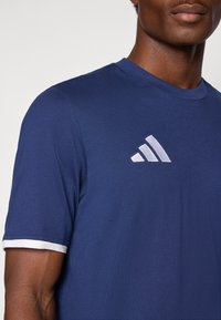 Μπλούζα τζιν κοντομάνικη σε ναυτικό μπλε χρώμα, κατασκευασμένη από μαλακό ύφασμα, με λευκό περίγραμμα στα μανίκια και λευκό λογότυπο Adidas στο στήθος.